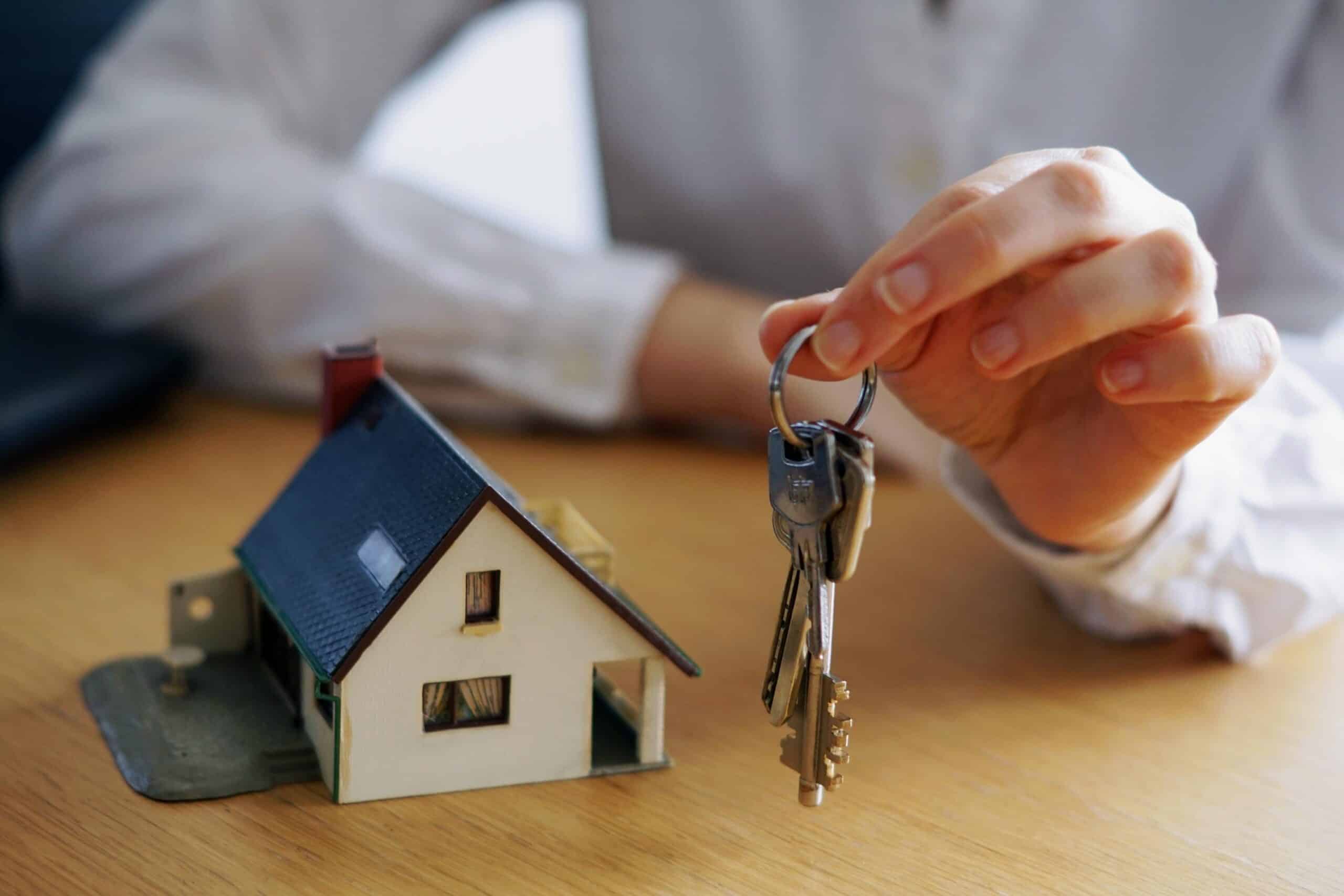 Hand holding house keys beside a model home, symbolizing a home purchase and new homeownership.